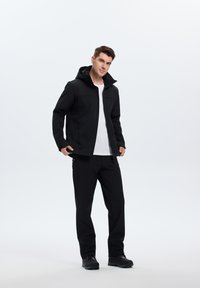 Veste softshell noire avec capuche et poches zippées, associée à un pantalon noir et des chaussures foncées, sur un fond léger uni.
