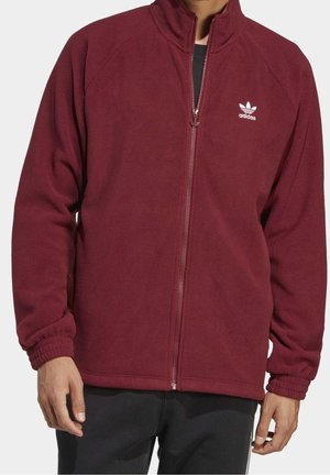Man wearing zipped maroon fleece jacket with elastic cuffs and white Adidas logo on left chest, paired with black pants.