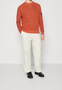 Orange ribbad tröja med rund halsringning och långa ärmar, ihop med vita cargo-byxor med sidofickor, och svarta skor.