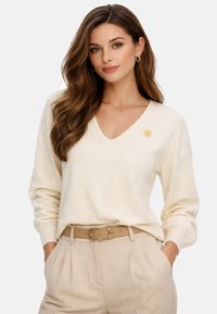 Femme aux cheveux bruns ondulés portant un pull à col en V crème avec un emblème de soleil et un pantalon beige plissé avec une ceinture marron clair, les mains dans les poches.