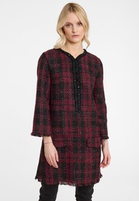 Rot-schwarz kariertes Tunika mit strukturiertem Finish, mit einem Fransenabschluss, langen Ärmeln und einer geknöpften Frontschließe.