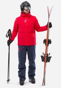 Veste de ski rouge avec poches zippées, pantalon de ski bleu marine avec accents rouges, casque noir, lunettes de ski, bâtons de ski et skis avec détails orange.