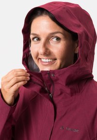 Giacca impermeabile bordeaux con cappuccio; presenta una chiusura con zip, logo visibile e un tessuto testurizzato. Mani che regolano la zip.