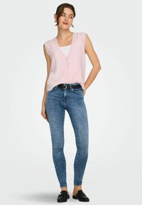 Jeune femme portant un gilet rose sans manches boutonné, un haut blanc, un jean skinny bleu avec une ceinture noire, et des chaussures noires sans lacets, debout avec la main dans la poche.