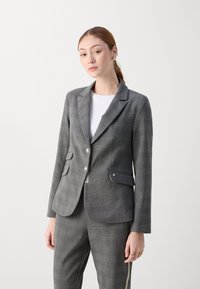 Grijze geruite blazer met een enkele rij knopen, notch revers, twee voorzakken en metalen knopen. Draagt over een witte top.