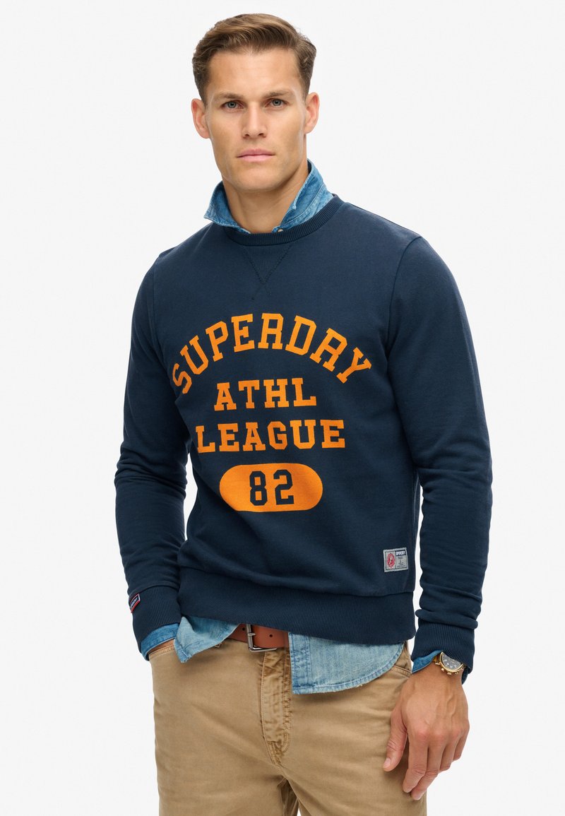 Sudadera azul marino con el texto naranja "SUPERDAY ATHL LEAGUE 82." Presenta una textura de mezcla de algodón, superpuesta sobre una camisa de mezclilla de color azul claro.