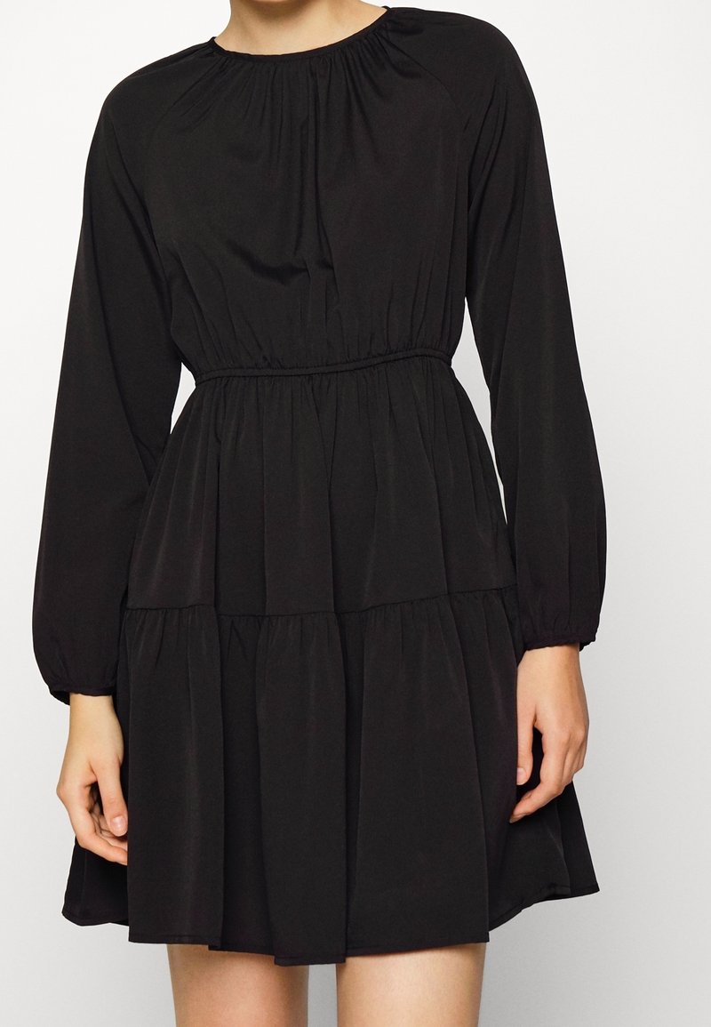 Femme portant une robe noire à manches longues avec une taille froncée et une jupe à volants, debout devant un fond blanc uni.