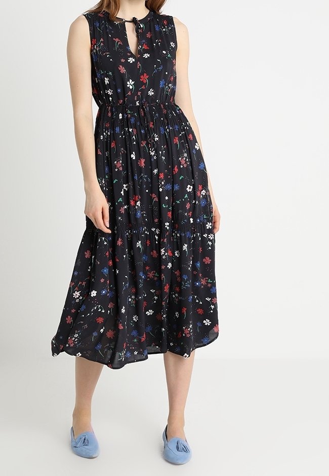 Vestido midi negro sin mangas con estampado floral multicolor, cintura fruncida, escote en forma de ojo de cerradura, combinado con zapatos planos celestes con borlas.