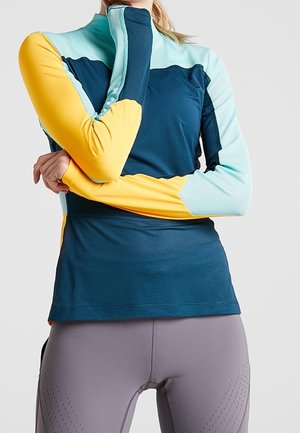 Frau trägt ein mehrfarbiges langärmliges Sportoberteil mit Daumenlöchern, kombiniert mit enganliegenden grauen Leggings.