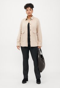 Beige suede shirt jacket with a collar, front button closure, and two large flap pockets. Black pants and a large black tote bag.
