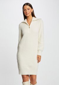 Abito maglione color crema realizzato in materiale peloso, con colletto a mezza zip e maniche lunghe, dalla vestibilità ampia e orlo svasato.