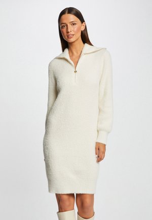 Abito maglione color crema realizzato in materiale peloso, con colletto a mezza zip e maniche lunghe, dalla vestibilità ampia e orlo svasato.
