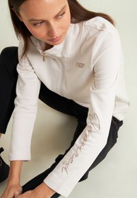 Donna seduta sul pavimento con una giacca bianca con zip con "FREDY" e testo scritto sulla manica, abbinata a pantaloni neri e sneakers nere.