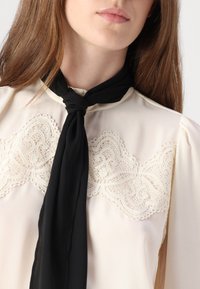Ivory bluse med blonderdetaljer ved halsen og sort bindebånd. Glatt tekstur, lange ermer og design med høy krage.