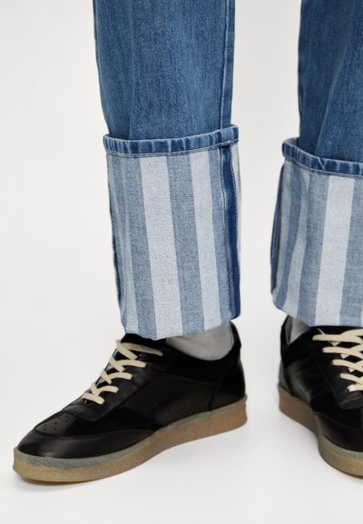 Zapatillas negras con suelas claras y cordones beige, combinadas con vaqueros de mezclilla azul que tienen un dobladillo a rayas en azul claro y oscuro.