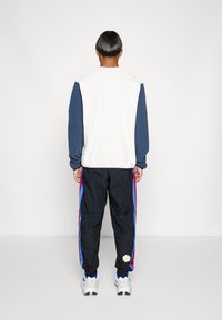 Vit och marinblå sweatshirt i kombination med svarta joggingsbyxor med blå och lila sidostreck samt en blommig applikation. Skorna är vita sneakers.