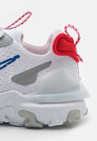 Zapatilla blanca con un upper texturizado, acentos grises, tirador rojo en el talón y una suela translúcida con el logo "Nike React".