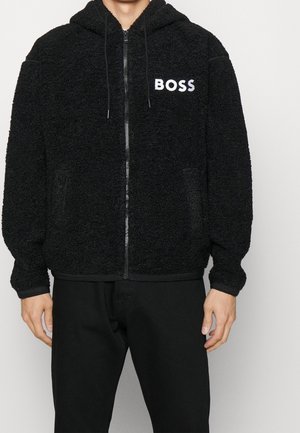 Mężczyzna ubrany w czarną, teksturowaną bluzę na zamek z logo "BOSS" na lewej piersi oraz czarne spodnie na jednolitym tle.