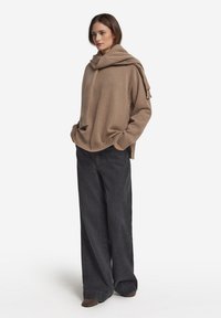 Khaki, oversize'owy sweter z prążkowanym wykończeniem i detalem w formie szalika, zestawiony z luźnymi ciemnoszarymi dżinsami oraz brązowymi butami. Prosty, teksturowany wzór.