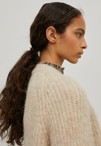 Beige gebreide trui met een textuurpatroon en een gerimpelde kraag. Model heeft lang, krullend bruin haar dat in een paardenstaart is gestyled.