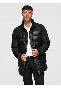 Hombre con chaqueta acolchada negra sobre camisa blanca y pantalones negros, ajustando la cremallera de la chaqueta, con tatuajes visibles en las manos, frente a un fondo sencillo.