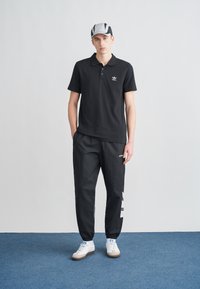 Mustvalge väikese logoga must poloülikond, millel on valgete triipudega mustad vabaajarõivad. Modell kannab halli nokamütsi ja valgeid tenniseid.