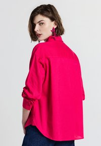 Donna con capelli corti castani che indossa una maglietta a maniche lunghe fucsia brillante, grandi orecchini floreali rosa e pantaloni scuri, che guarda oltre la spalla.