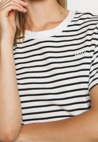 Nainen, joka käyttää valkoista ja mustaa vaakaraitapaita, jossa on pieni kirjailtu Levi's-logo, käsi lähellä kasvoja, kuva on rajattu hartioista.