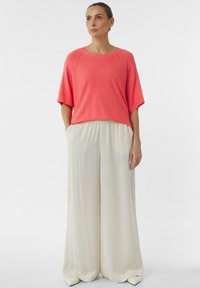 Korallowa bluzka z krótkim rękawem o okrągłym dekolcie, zestawiona z szerokimi beżowymi spodniami z bocznymi kieszeniami i teksturowanym wykończeniem.