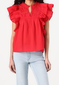 Haut en coton rouge avec manches volantées et encolure en goutte d'eau, présentant des détails smockés et une coupe ample. Associé à un jean bleu clair.