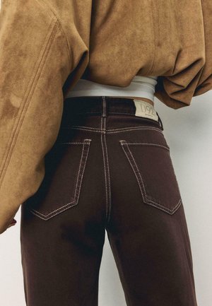 Persoon draagt donkerbruine spijkerbroek met witte stiksel en een tan suède jas, waarbij het achterste taillegebied en de zakken zichtbaar zijn.