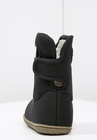 Botín de tela negro hasta el tobillo con un forro blanco de felpa, que cuenta con una correa de Velcro, suela texturizada y diseño de punta redondeada.