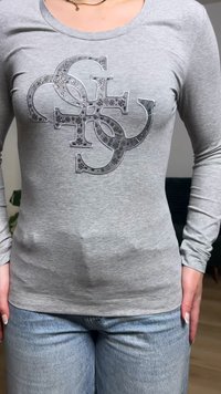 Kvinde iført en grå langærmet skjorte med pyntede overlappende bogstaver og lyseblå jeans, stående indendørs.