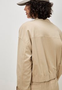 Vrouw met krullend donker haar, gekleed in een beige jas, bijpassende broek en een geruite pet, zijkant kijkend tegen een effen achtergrond.
