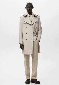 Beżowy płaszcz trencz z dwurzędowym zapięciem, dużym kołnierzem, przednimi kieszeniami i guzikami w tonacji, zestawiony z jasnobeżowymi spodniami i ciemnymi butami.