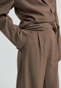 Personne portant un pantalon marron taille haute à jambes larges avec une chemise marron à manches longues assortie rentrée à l'intérieur, main posée sur la hanche.