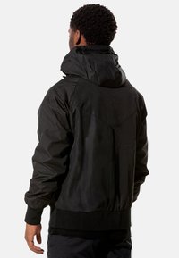 Schwarze Jacke mit Kapuze, aus strukturiertem Stoff, mit elastischen Bündchen und einem Gummizug am Saum sowie einem einzigartigen Nahtdesign auf dem Rücken.