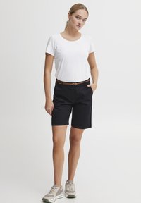 Shorts noirs avec un léger éclat, ceinture marron et t-shirt à col rond blanc. Le modèle porte des baskets gris clair. Tenue simple et décontractée.