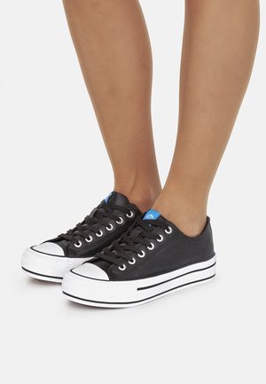 Zapatillas de lona negras con suelas de goma blanca y protectores de punta blancos, cordones negros y un acento azul cerca de la lengüeta, con un diseño de perfil bajo.