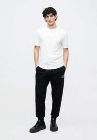 Λευκό κοντομάνικο μπλουζάκι με χρυσό λογότυπο, συνδυασμένο με μαύρο στενό παντελόνι φούτερ με λεπτομέρεια λογότυπου και μαύρα αθλητικά παπούτσια.