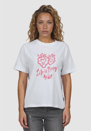 Vrouw met krullend haar die een wit T-shirt draagt met roze cartoonbessen en de tekst "Life is Berry nice," gecombineerd met grijze jeans tegen een effen achtergrond.