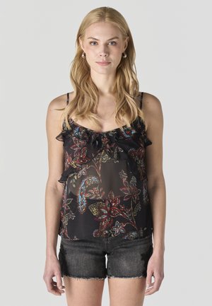 Mujer rubia con una blusa sin mangas con volantes y estampado floral en negro, y pantalones cortos de mezclilla negros, de pie frente a un fondo gris claro.