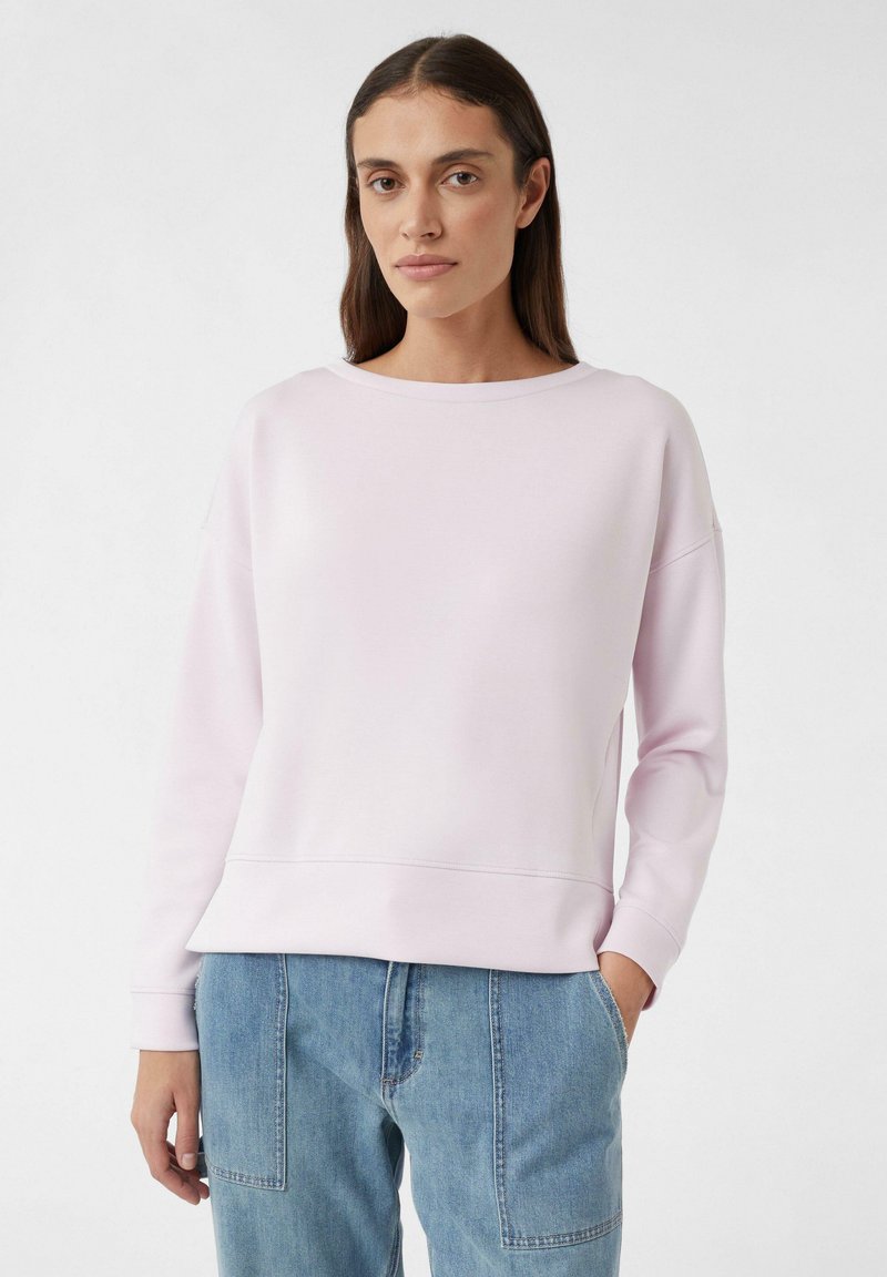 Jasnoróżowy sweter wykonany z miękkiego materiału, z okrągłym dekoltem, opadającymi ramionami i ściągaczami na rękawach, zestawiony z niebieskimi dżinsami.