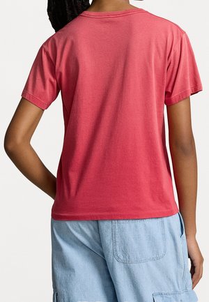 Osoba ubrana w gładki koralowy t-shirt z krótkim rękawem oraz jasnoniebieskie dżinsowe szorty, pokazana od tyłu na białym tle.