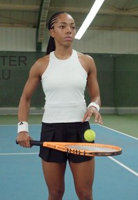Nainen tennispelaaja valkoisessa hihattomassa paidassa ja mustissa shortseissa pitää oranssia tennismailaa, jossa on keltainen tennipallo, kentän edessä.
