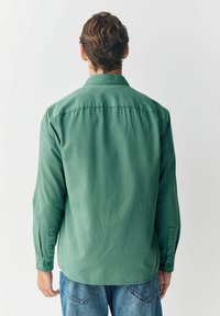 Grüne Langarmshirt aus weichem Baumwollstoff, mit Knopfmanschette, spitzem Kragen und abgerundetem Saum. Getragen mit Jeans.