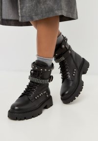 Botas de cuero negras hasta el tobillo con una suela gruesa, adornadas con tachuelas metálicas y una banda de rhinestone, con cordones y hebillas laterales.