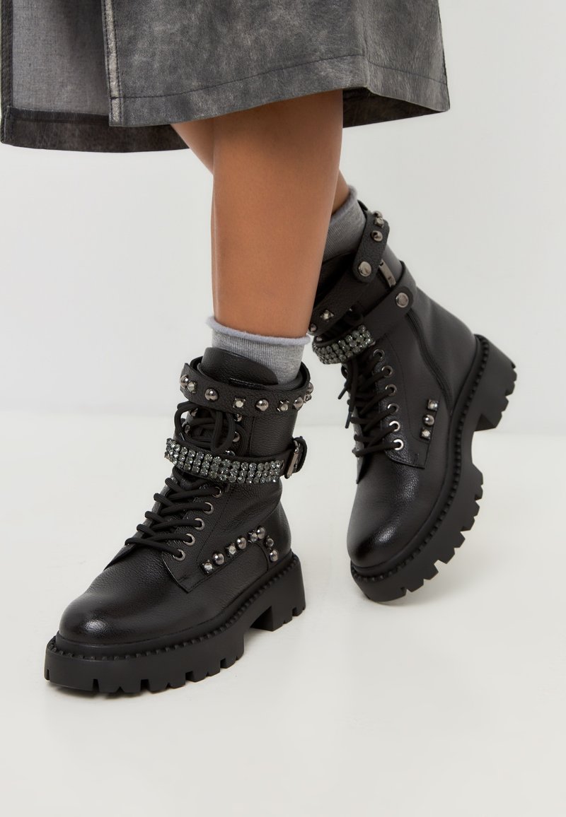 Botas de cuero negras hasta el tobillo con una suela gruesa, adornadas con tachuelas metálicas y una banda de rhinestone, con cordones y hebillas laterales.
