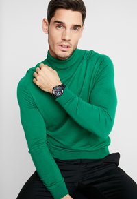 Groene coltrui van zachte stof, nauwsluitende pasvorm, met ribbelmanchetten en -zoom. Een zwart polshorloge met een ronde wijzerplaat wordt om de pols gedragen.