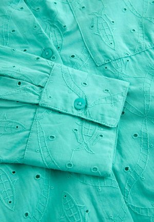 Turkis bomuldsbluse med broderede fisk og svungne mønstre, udstyret med knapmanchetter og små udskæringer.